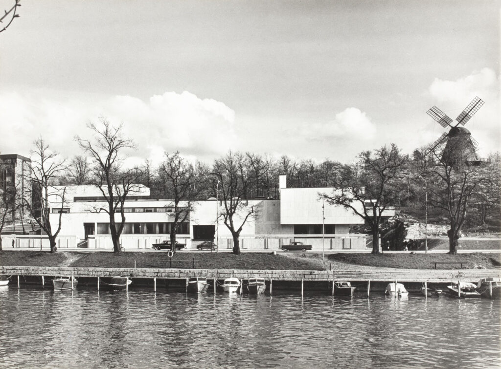 Mustavalkoinen valokuva jossa valkoinen, kulmikas museorakennus näkyy kauniisti joen varrella.